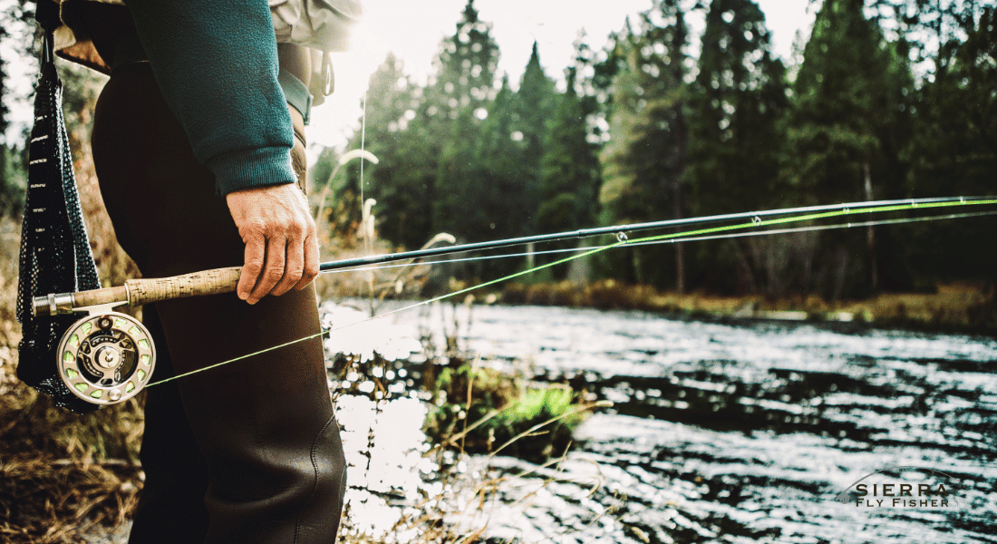 A man stands outdoors, confidently holding a fly fishing rod and reel, ready for a day of fishing.