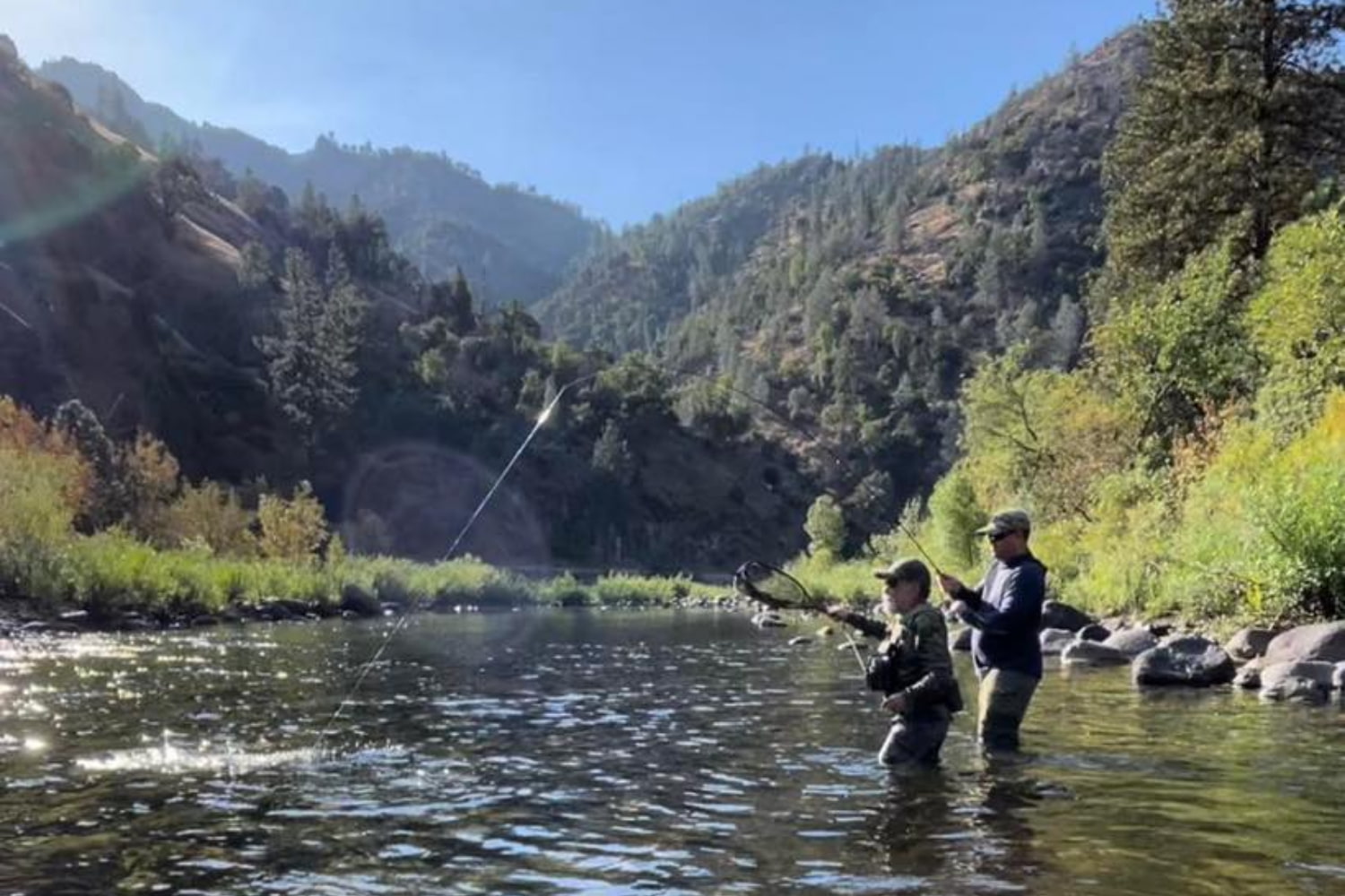 Fly fishing guide instructing a guest while wading in a clear High Sierra river with granite peaks and morning sun in the background